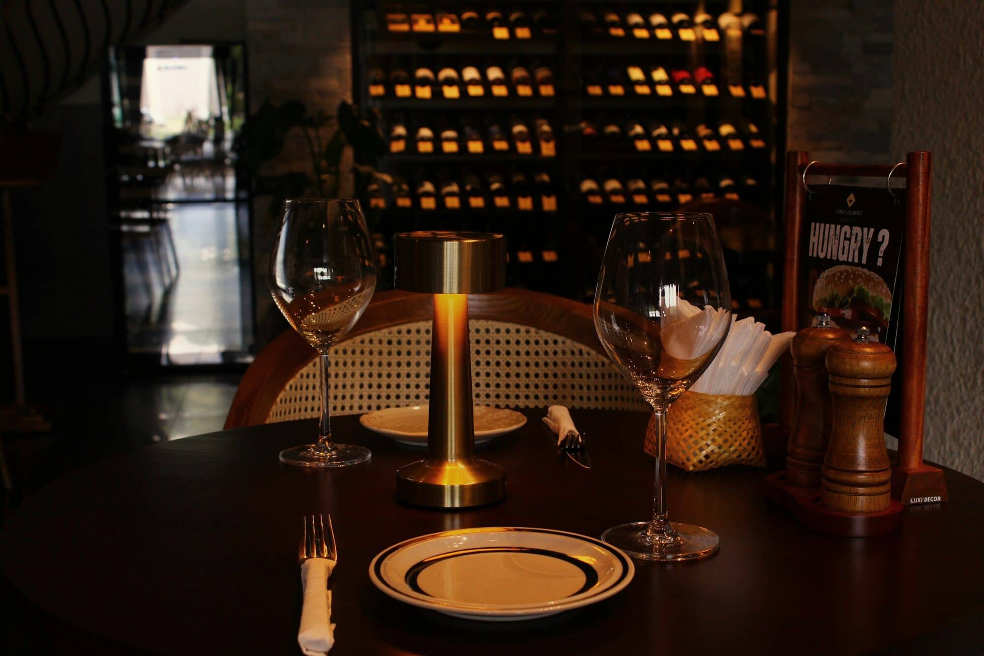 A table set for dinner under a small brass lamp, a wall of wine behind it